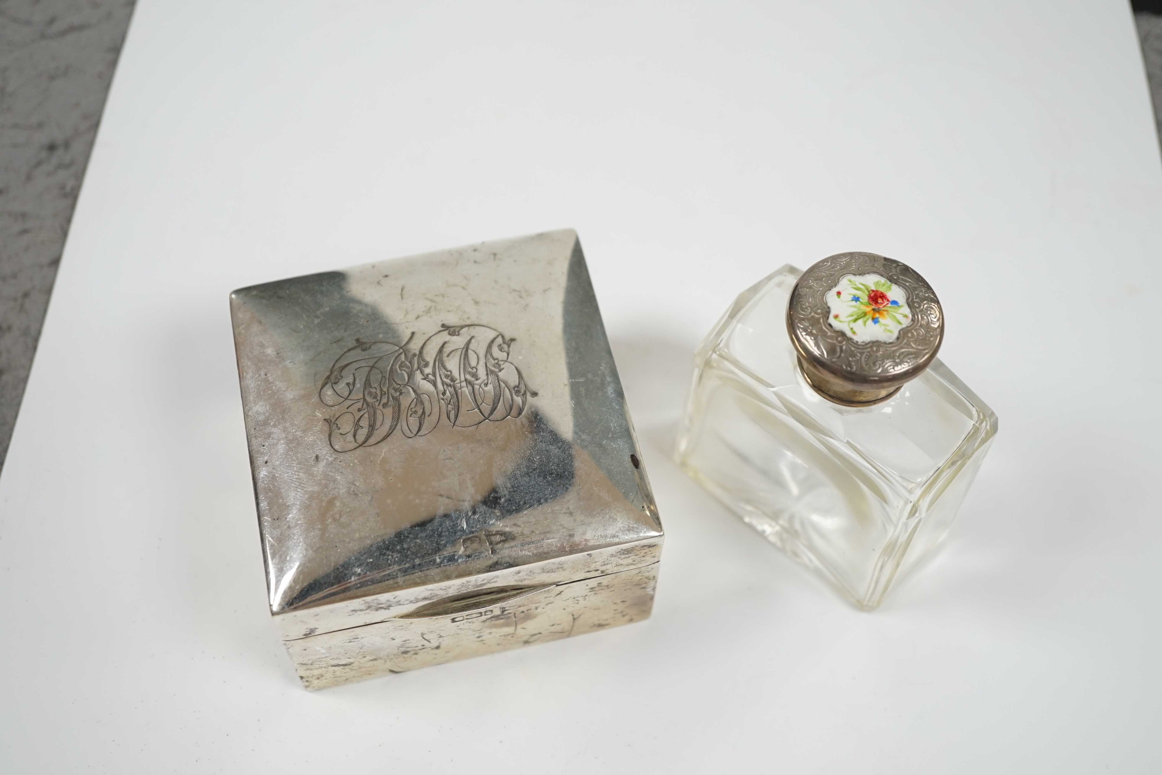 An Edwardian silver mounted cigarette box, marks rubbed, 90mm, together with a white metal and enamel topped glass scent bottle. Condition - poor